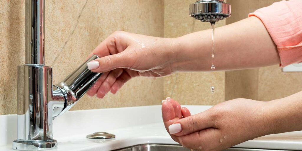 A person turns a kitchen faucet handle as only a weak trickle of water drips out, showing low water pressure.