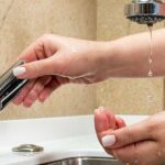 A person turns a kitchen faucet handle as only a weak trickle of water drips out, showing low water pressure.