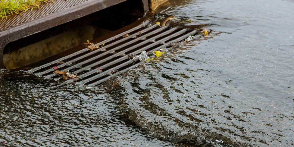 Stormwater runoff flows rapidly over a metal street storm drain grate, causing localized pooling during a heavy rainstorm event.