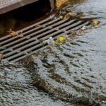 Stormwater runoff flows rapidly over a metal street storm drain grate, causing localized pooling during a heavy rainstorm event.