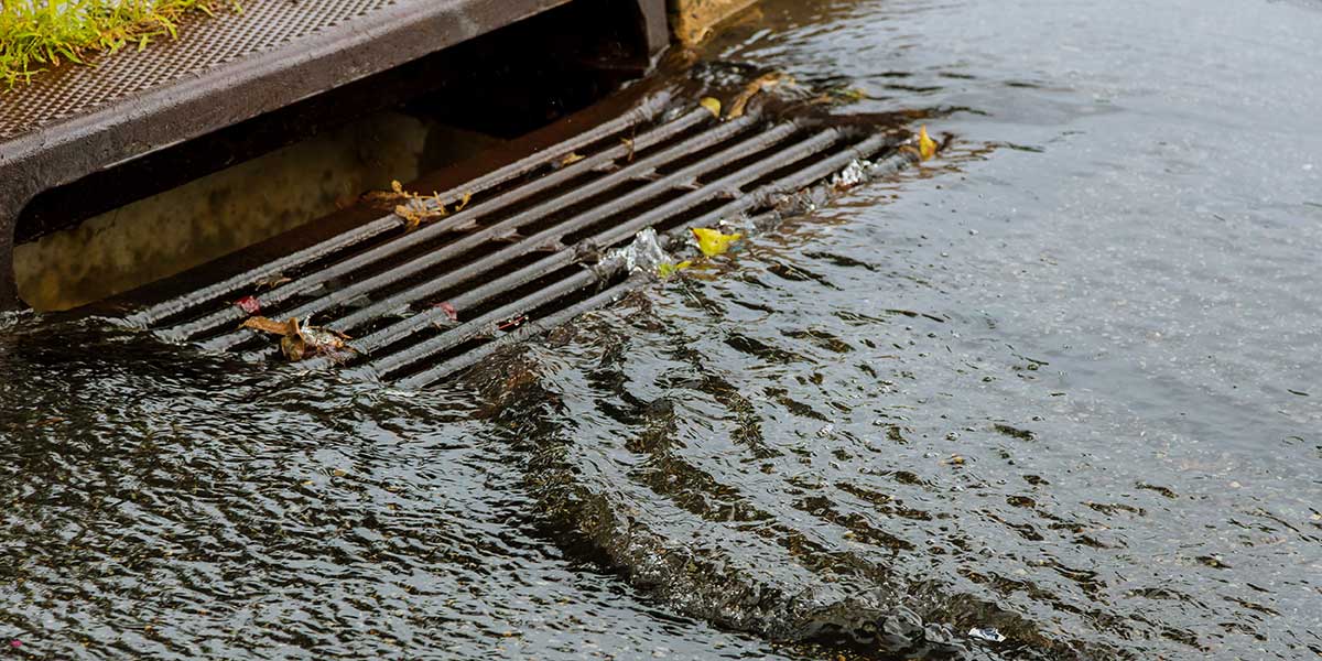 Stormwater runoff flows rapidly over a metal street storm drain grate, causing localized pooling during a heavy rainstorm event.