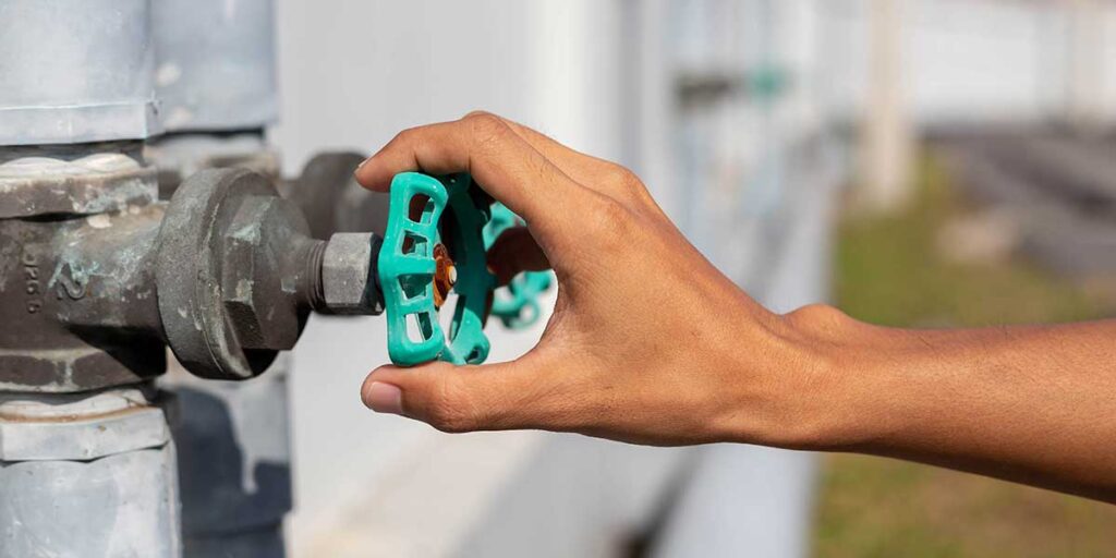 A person's hand turning a green wheel handle on an aged outdoor metal water shutoff valve mounted to pipes.