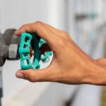 A person's hand turning a green wheel handle on an aged outdoor metal water shutoff valve mounted to pipes.