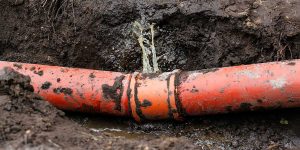 A cracked underground sewer pipe leaks wastewater into a muddy trench, showing visible damage that requires repair.
