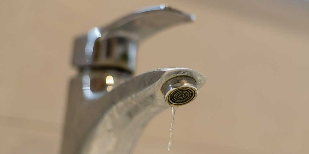 A close-up view of a chrome bathroom faucet slowly dripping a stream of water, highlighting household water waste and leaks.