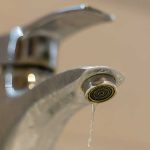 A close-up view of a chrome bathroom faucet slowly dripping a stream of water, highlighting household water waste and leaks.