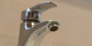 A close-up view of a chrome bathroom faucet slowly dripping a stream of water, highlighting household water waste and leaks.